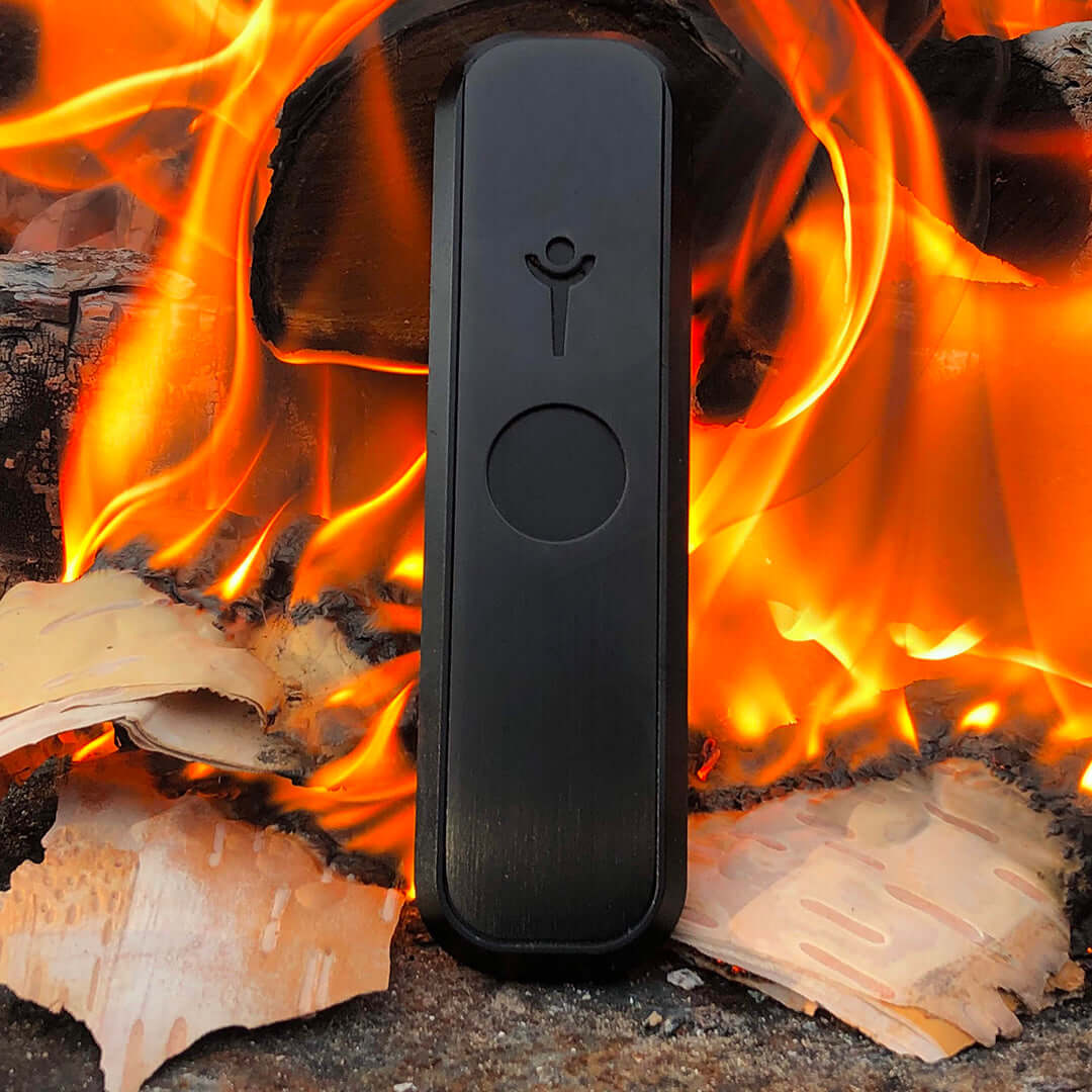 Genius Pipe Mini Black on surface with burning fire in background β warm lifestyle photo.